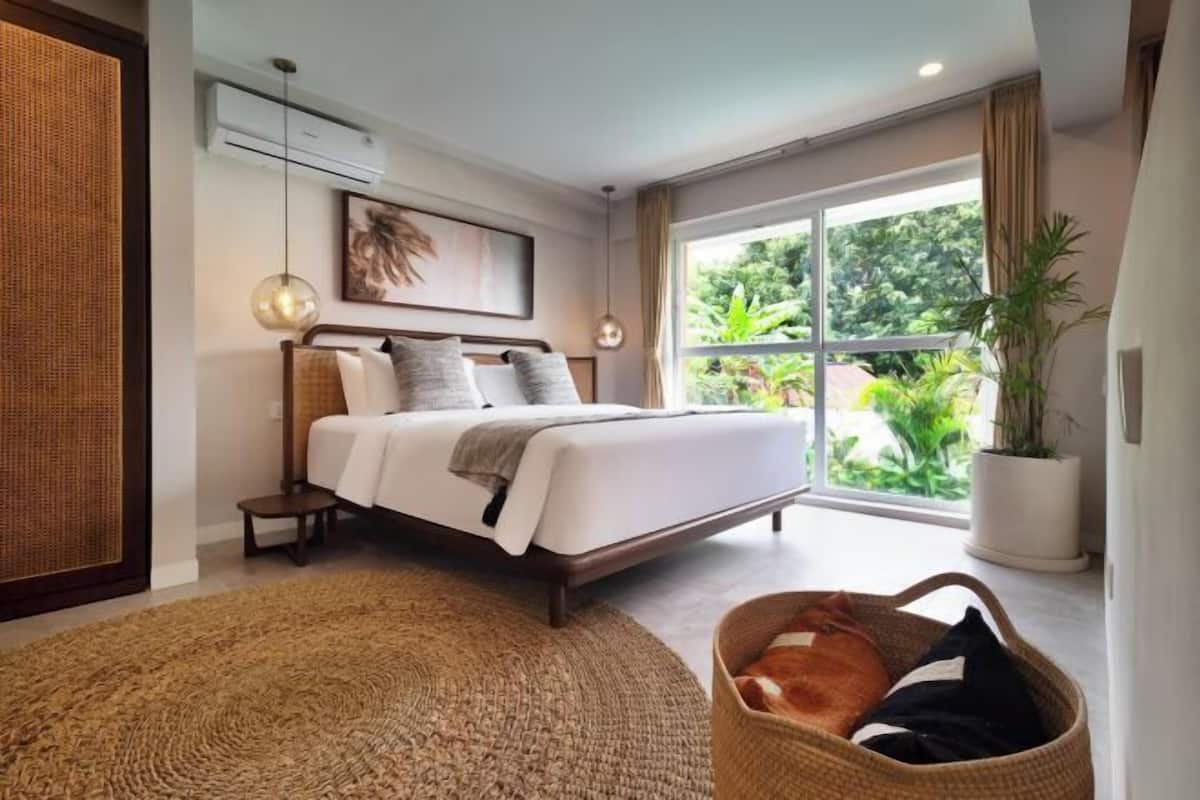 A modern bedroom with a large bed, a small bedside table, a hanging light fixture, a large window with a view of greenery, and a potted plant.