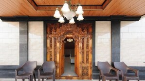 Une porte d’entrée en bois richement décorée, surmontée d’un lustre, avec deux fauteuils en osier et un sol carrelé.