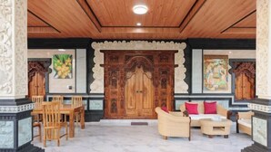 Un hall d’entrée avec un plafond en bois, une grande porte en bois richement décorée, un canapé avec des coussins et un coin repas avec des chaises.