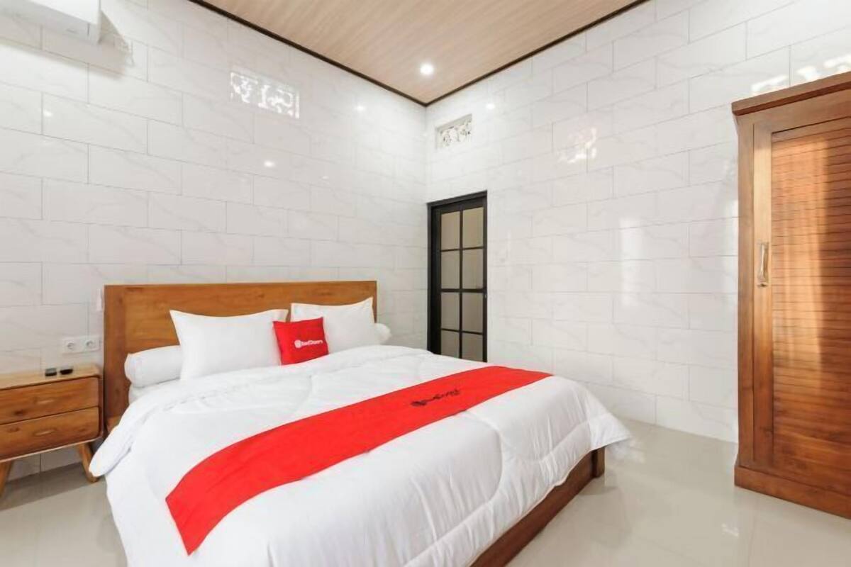 Habitación de hotel con cabecera de madera, cama con ropa de cama blanca y un detalle rojo, armario de madera y pared con azulejos blancos.