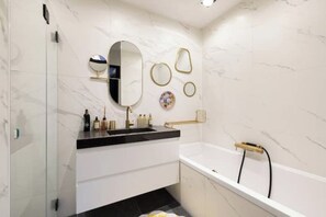 A modern bathroom with a marble wall, a large mirror, a bathtub, and a vanity with a black countertop.