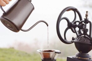 A hand pouring liquid from a metal pitcher into a coffee dripper over a coffee machine. - Thenor (Samoeng)