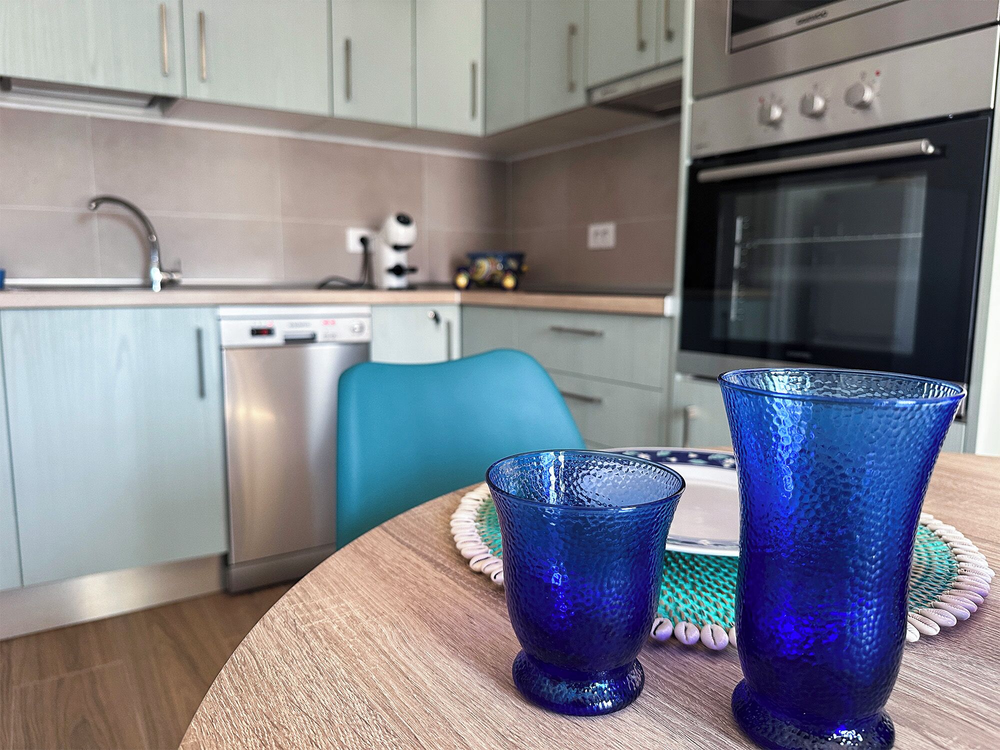 Une cuisine moderne avec des meubles de couleur claire, un lave-vaisselle et un four. On y trouve une chaise bleue et deux verres bleus sur une table en bois.