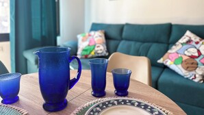 Una sala de estar con una mesa de centro de madera, una jarra azul y cuatro vasos azules. Se observa un sofá verde con almohadones de colores al fondo.