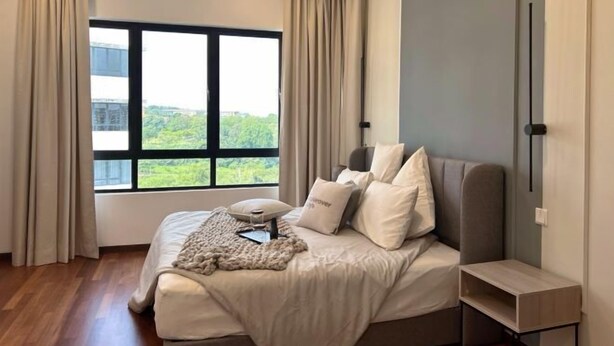 A bedroom with a large window, a bed with multiple pillows, a bedside table, and a view of greenery outside.