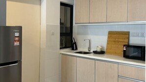 A modern kitchen with light wood cabinets, a stainless steel refrigerator, a black microwave, and a wooden cutting board on the countertop.
