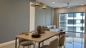 A modern dining area with a wooden table, chairs, and pendant lights. There is a sofa and a large window in the background.