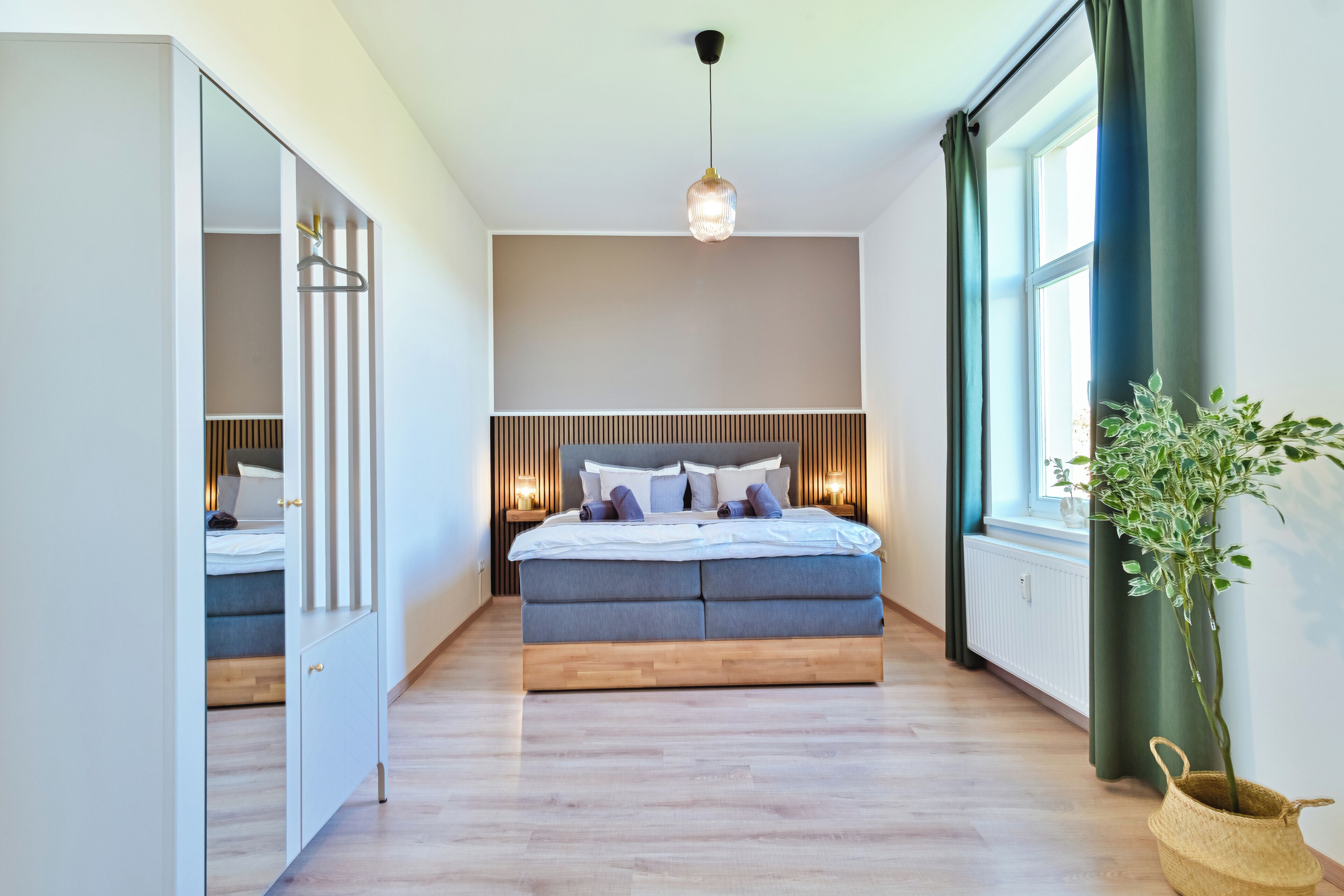 Une chambre à coucher moderne avec un lit, des lampes de chevet, une plante en pot et une vue sur l’extérieur.