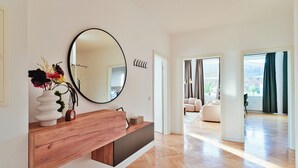 Una sala moderna con una consola de madera, un espejo redondo y un florero con flores. Se ve un pasillo que conduce a otra habitación con vistas al exterior.