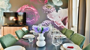 Un comedor con una mesa moderna preparada para seis personas, un jarrón blanco con flores y un televisor sobre un mueble de madera.