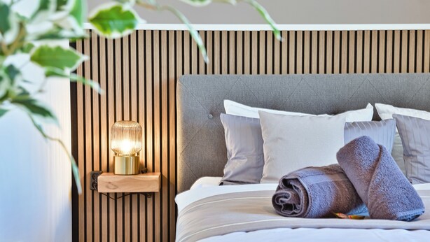 Una cama bien hecha con cabecera gris, una lámpara de noche y toallas enrolladas.
