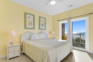 Une chambre à coucher avec un lit, deux tables de chevet avec des lampes, une fenêtre donnant sur la plage et un ventilateur de plafond.