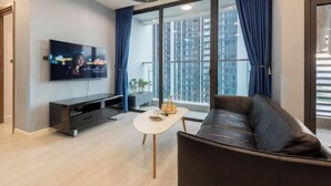 A modern living room with a flat-screen TV, a leather sofa, a small white table, and large windows with blue curtains. - Vinhome Metropolis Luxury Apartment - Tiramisu House (Hanoi)