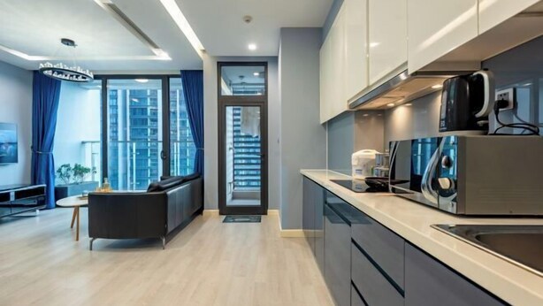 A modern kitchen with stainless steel appliances, a black sofa, and a view of a cityscape through large windows. - Vinhome Metropolis Luxury Apartment - Tiramisu House (Hanoi)
