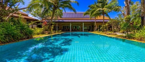 A swimming pool with a tiled surface, surrounded by palm trees and greenery, with a building in the background.
