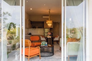 Un salon moderne avec une table basse ronde, deux fauteuils orange et une vue sur la cuisine et la salle à manger, grâce à une porte vitrée.