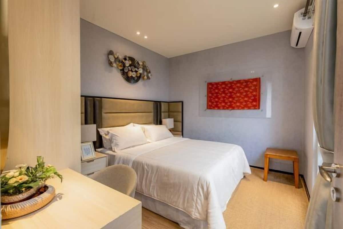 A hotel room with a bed, bedside table, chair, wall art, and a small stool.