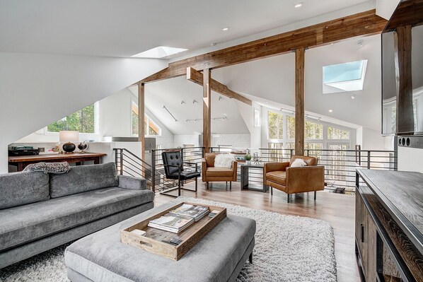 A spacious loft with a high ceiling, exposed wooden beams, a mezzanine level, and large windows providing natural light.