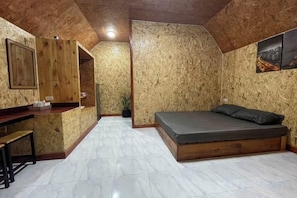 A room with a wooden ceiling, a bed with a mattress and pillows, a small table with chairs, and a wall-mounted shelf.
