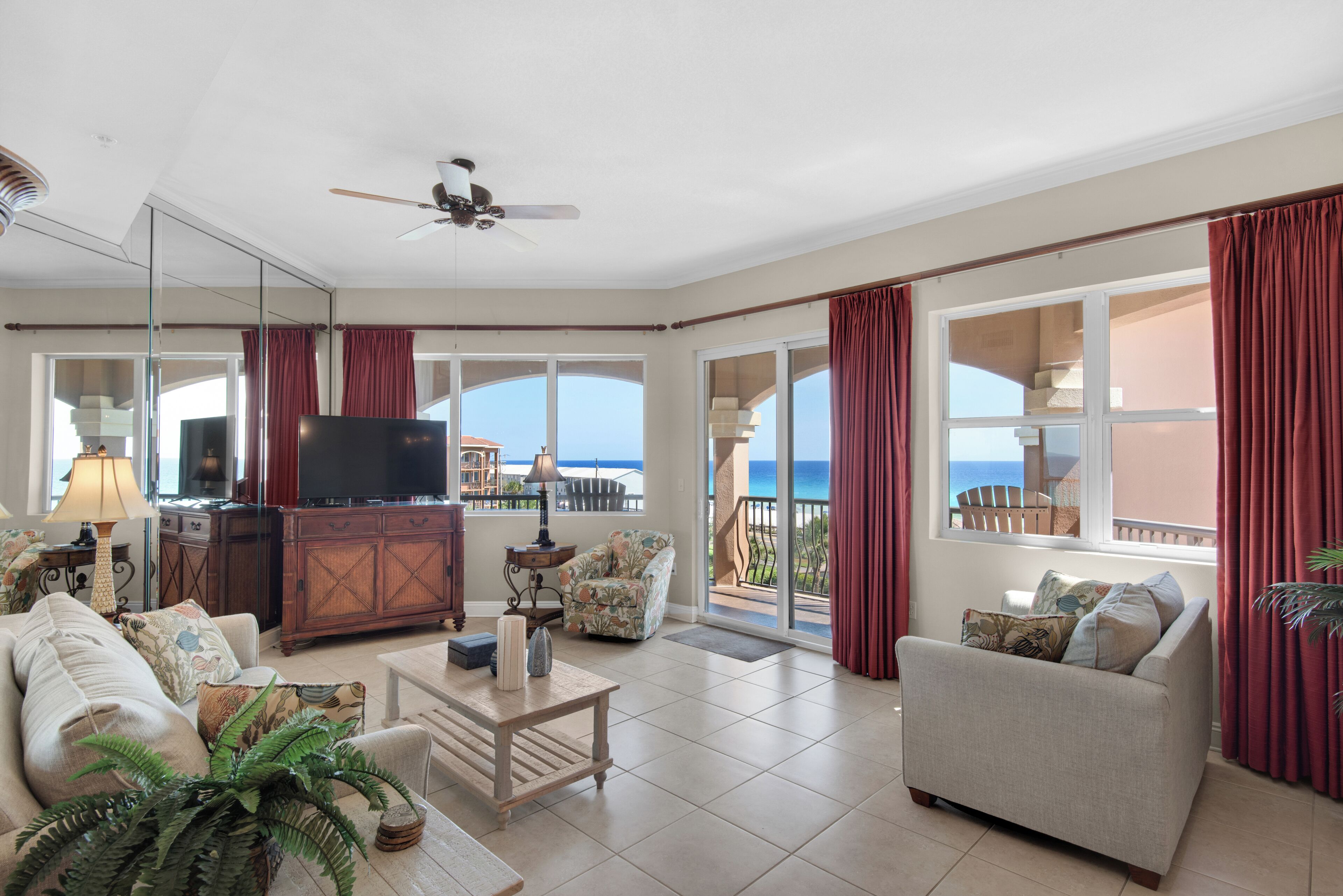 A spacious living room with a large TV, comfortable seating, a ceiling fan, and a view of the ocean through large windows.
