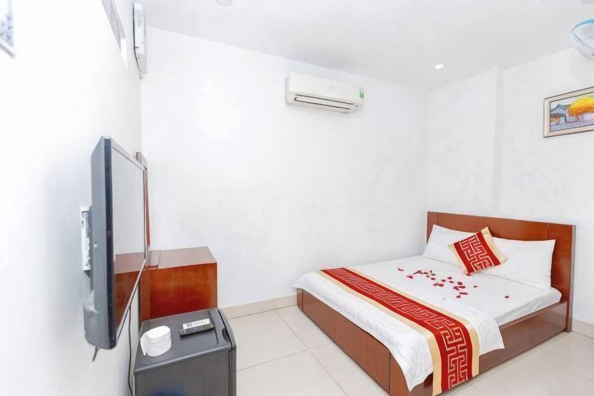 A hotel room with a bed, a TV, a bedside table, and an air conditioning unit.