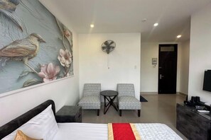 Une chambre d’hôtel avec un lit, deux chaises, une petite table, un ventilateur et un tableau au mur.