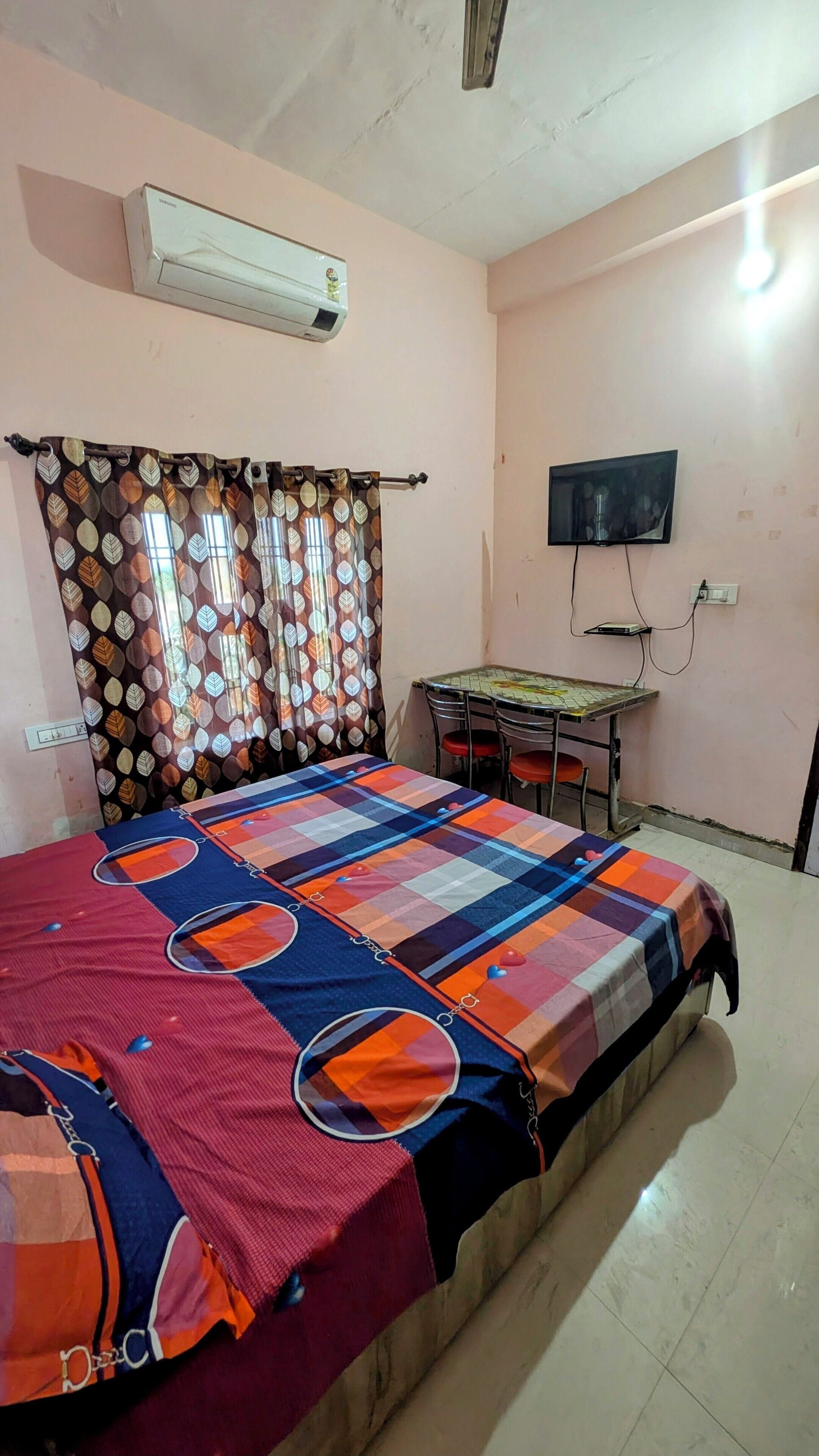 A bedroom with a bed, a table, a chair, an air conditioner, and a TV.