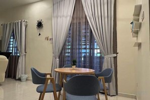 Uma sala de jantar com uma mesa redonda e quatro cadeiras, uma janela com cortinas e uma prateleira na parede com objetos.