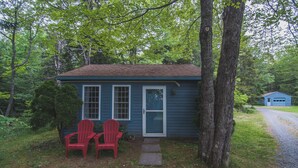 Eine kleine, blaue Holzhütte mit zwei roten Adirondack-Stühlen davor, ein Kiesweg führt zur Tür, und im Hintergrund steht eine blaue Garage.