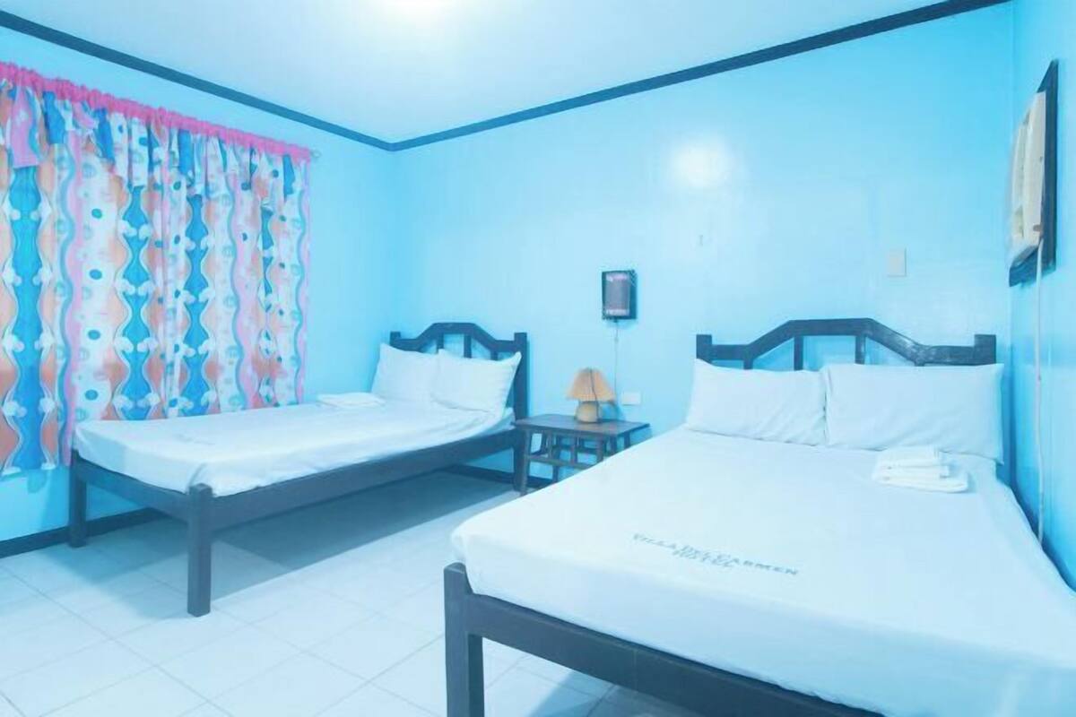 A room with two beds, a small table with a lamp, and a wall-mounted air conditioning unit.
