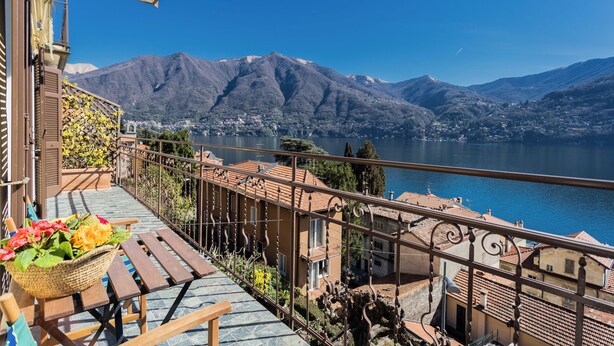 A balcony with a wicker basket of flowers, a wooden table, and a view of a lake with mountains in the background. - Lovely Apartment Overlooking the Lake (Carate Urio)