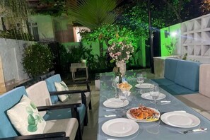 Un área de comedor al aire libre con una mesa preparada para dos personas, rodeada de asientos acolchados azules, un florero con flores y un fondo de follaje y una pared blanca.