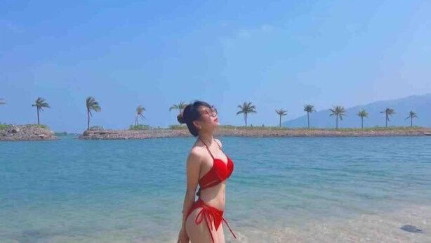 Une femme vêtue d’un bikini rouge se tient sur une plage, avec une eau cristalline et des palmiers en arrière-plan.
