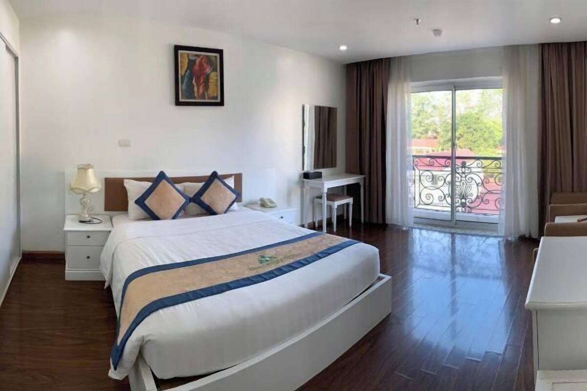 Quarto de hotel com cama, escrivaninha, cadeira, televisão e varanda com guarda-corpo.