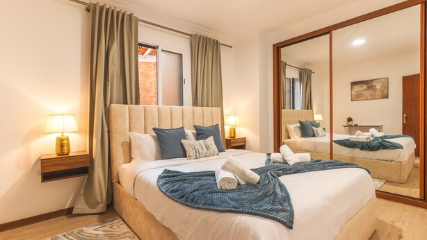Une chambre moderne avec un grand lit, deux tables de chevet avec des lampes, un miroir et une vue sur un mur de briques à travers une fenêtre.