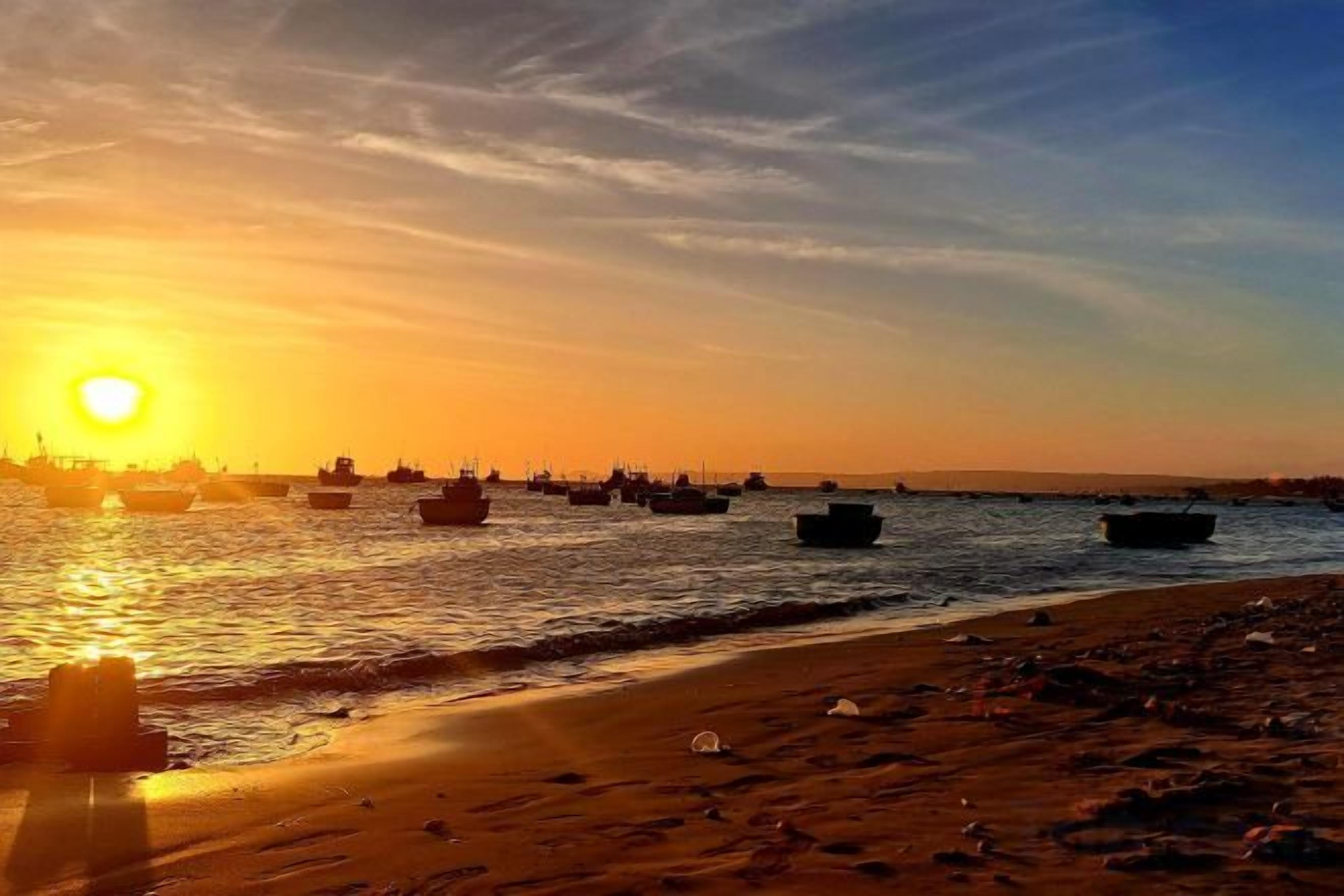 A beach with boats on the water and a sunset in the background.
