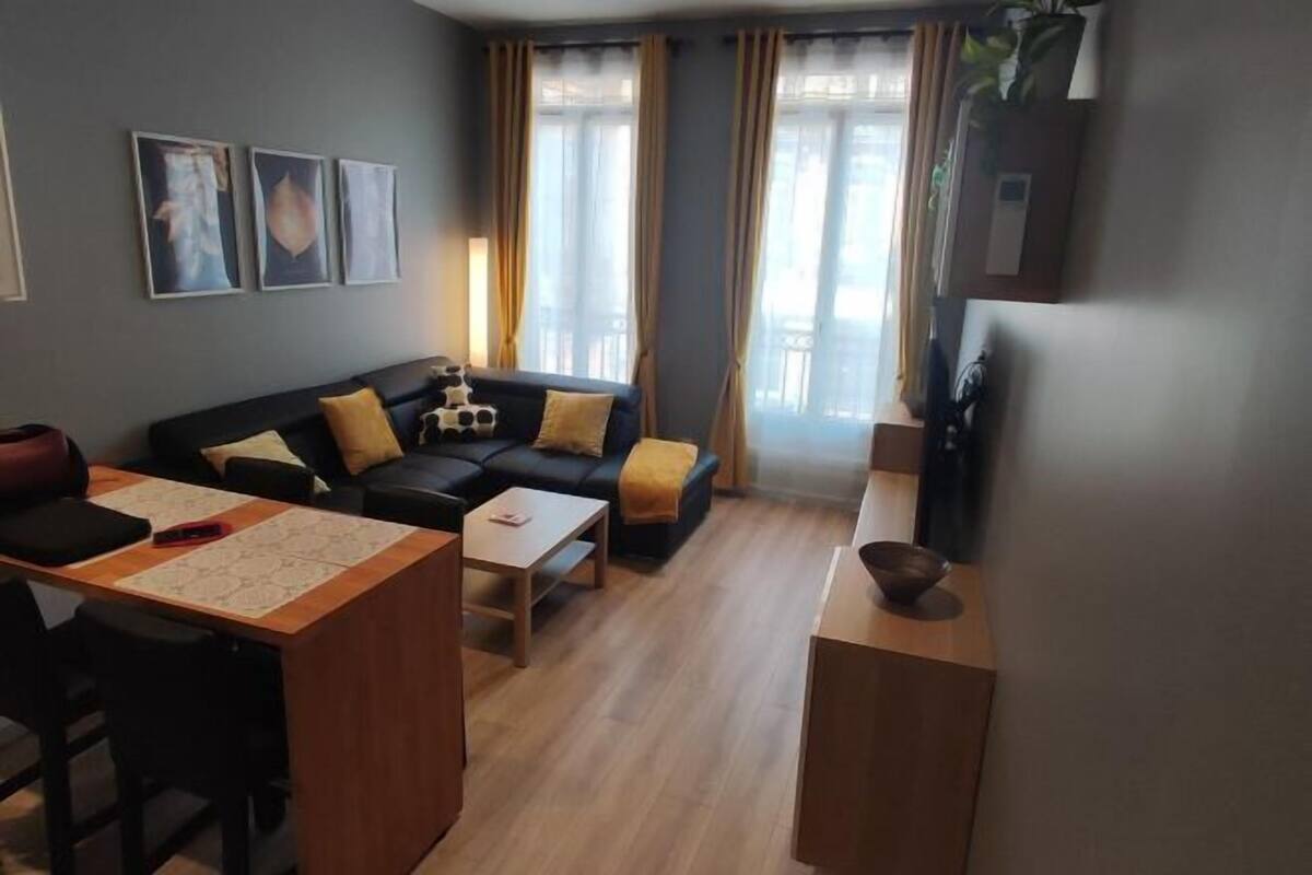 A living room with a sofa, coffee table, and a TV. There are curtains, a plant, and a wall-mounted picture.