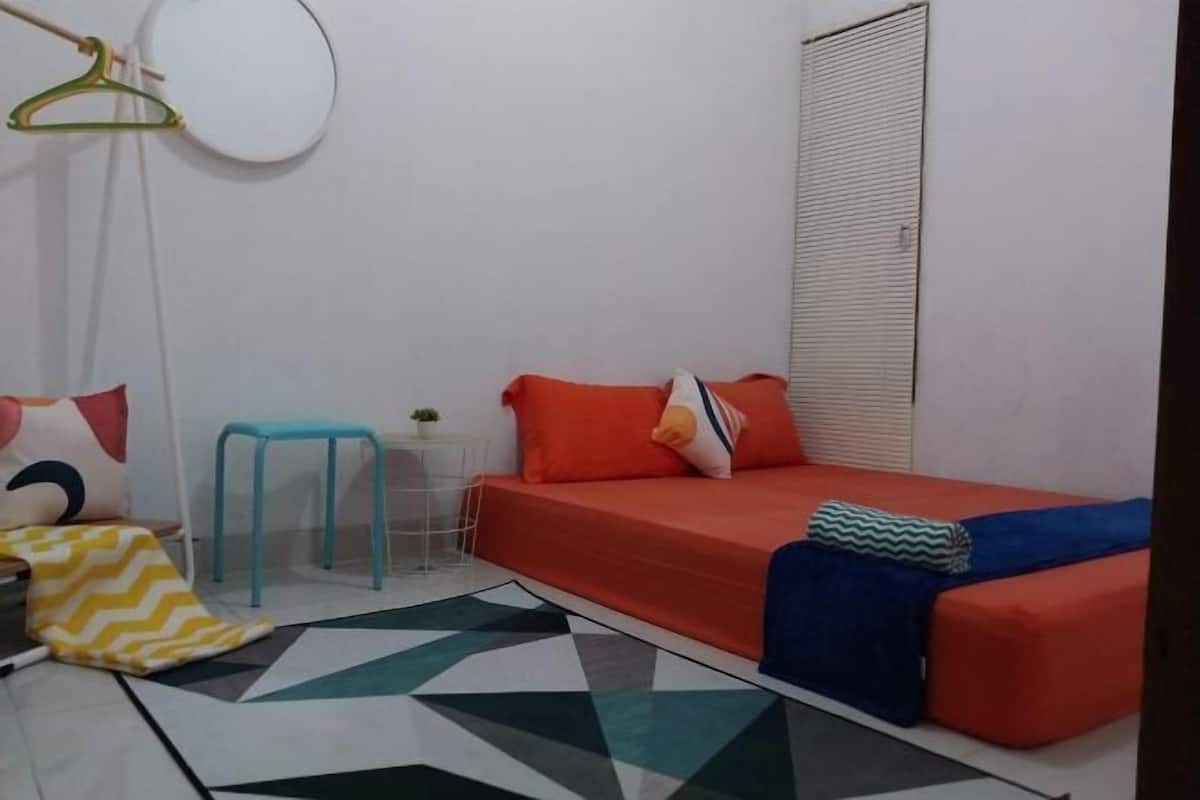 A small, single-bedroom room with a bed, a chair, a small table, and a hanging light fixture.