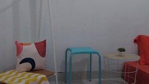 A small, neatly arranged room with a single bed, a blue stool, a small round table with a plant, and a red chair.