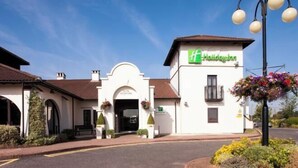 Un edificio blanco con un letrero verde de 'Holiday Inn', un acceso cubierto y una maceta con flores a la derecha.