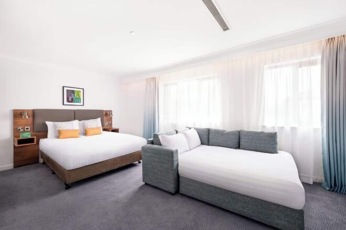 A hotel room with a double bed, a sofa bed, a wooden nightstand, and a framed picture on the wall.