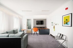 A modern hotel room with a grey sofa, a small dining table with an orange chair, a kitchenette, and a TV mounted on the wall. - Holiday Inn BIRMINGHAM - BROMSGROVE by IHG (Bromsgrove)