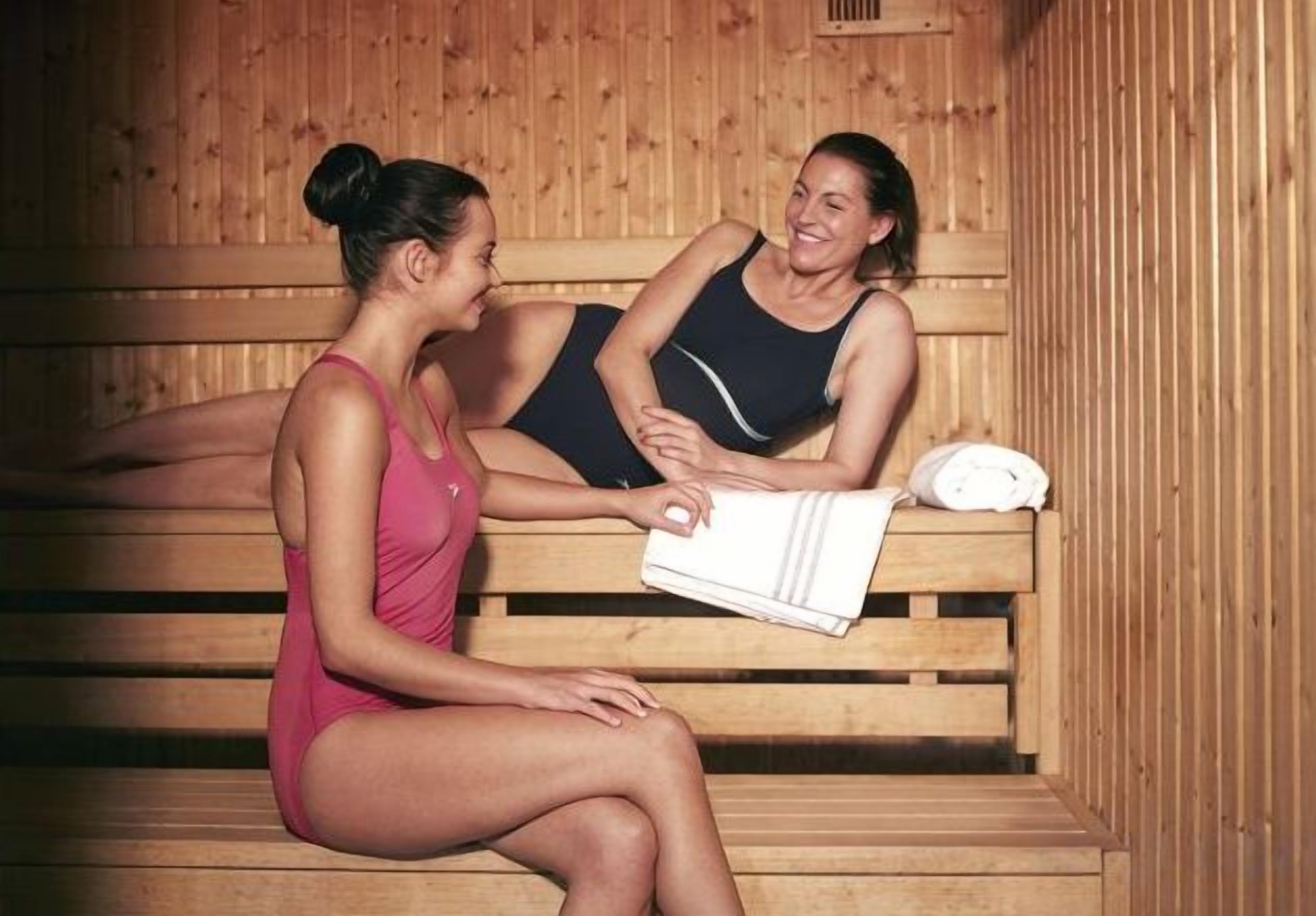 Two individuals seated on wooden benches in a sauna room with benches and a towel on the right.