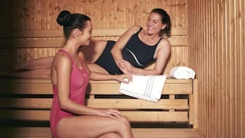 Two individuals seated on wooden benches in a sauna room with benches and a towel on the right.