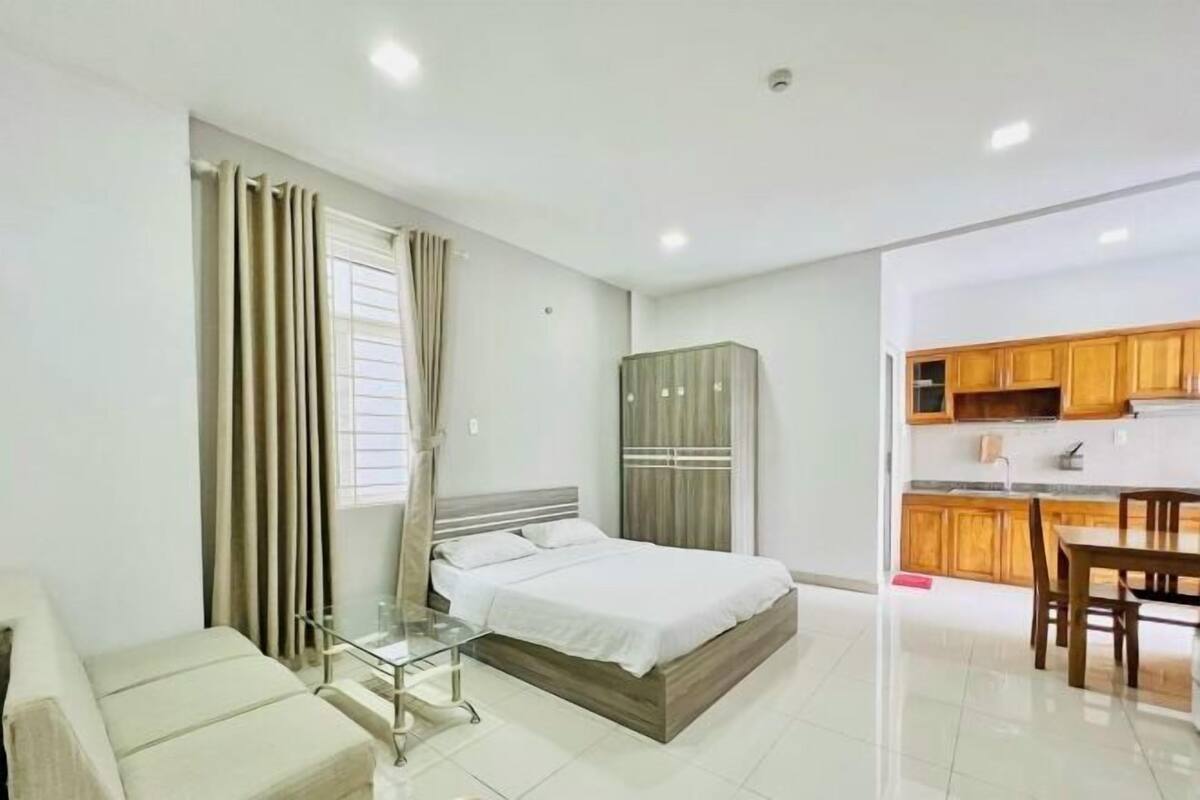 A bedroom with a bed, a glass table, a sofa, a wardrobe, and a kitchen area with wooden cabinets and a dining table.