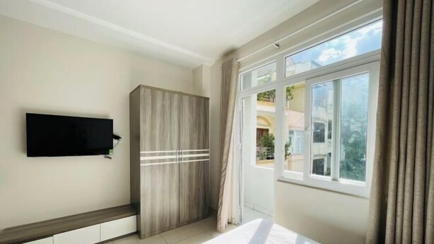 A modern bedroom with a large bed, a wardrobe, a flat-screen TV mounted on the wall, and a window with curtains. - EMERALD APARTMENT (Ho Chi Minh City)