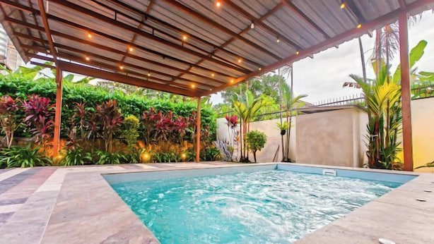 A swimming pool with a tiled deck, surrounded by lush greenery and a covered area with string lights. - JB Town Villa by RR JBcity (Johor Bahru)