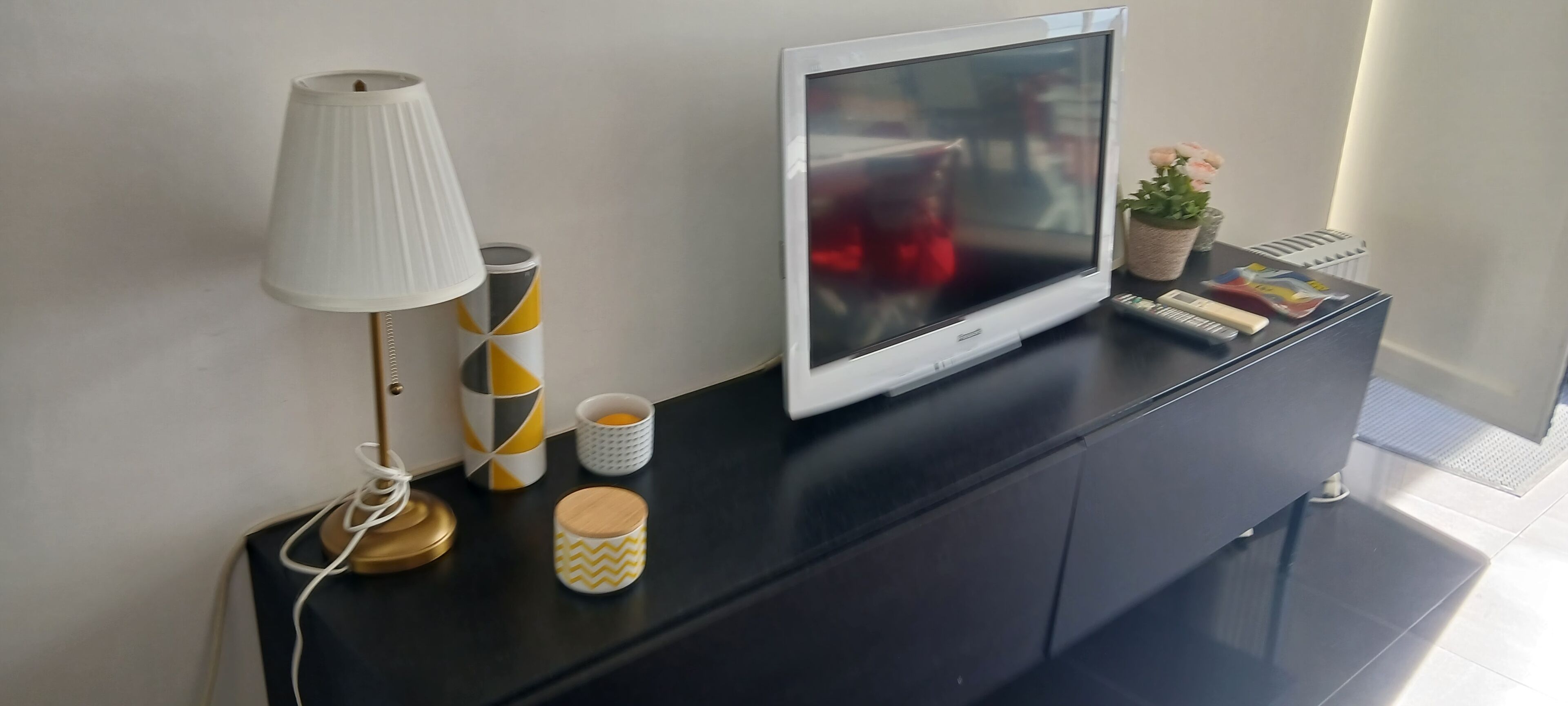 Ein modernes Wohnzimmer mit Fernseher auf einer schwarzen Unterhaltungsecke, einer Lampe, dekorativen Behältern und einer kleinen Pflanze.
