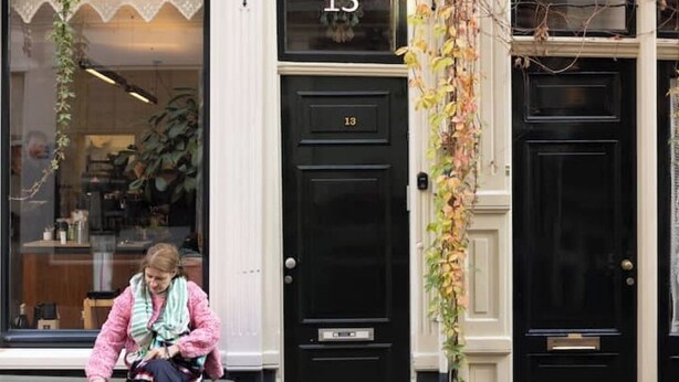 A person sitting on a bench outside a building with a black door marked with the number 13.