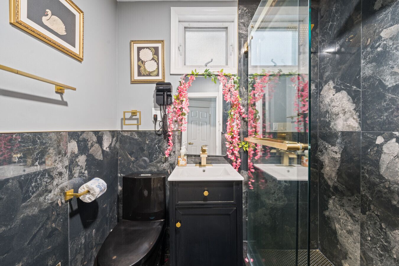 Un baño con un lavabo negro, un inodoro y una ducha. Hay cuadros en la pared y un espejo con una guirnalda de flores.
